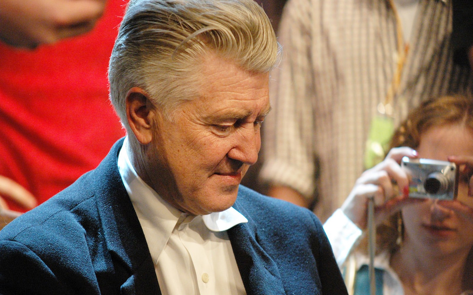 Grey-haired man in a navy blazer looks down as onlookers photograph him